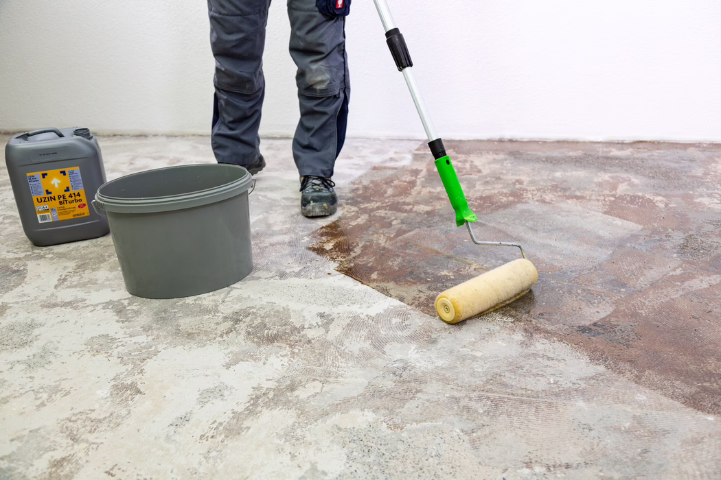 UZIN_Application Image_PE 414_9406_2018-11 On voit les jambes jusqu'aux pieds d'un ouvrier qui applique, à l'aide d'un rouleau à manche long, une couche brune de primaire ou d'adhésif sur un sol en béton brut. Un seau en plastique gris et un bidon gris portant l'étiquette "UZIN PE 414 Biturbo" sont posés à proximité contre un mur blanc. L'ouvrier porte un pantalon de travail gris et des chaussures de sécurité noires ; le sol montre des traces d'anciens adhésifs et du béton poncé pendant que le rouleau étale le produit pour préparer la sous-couche.