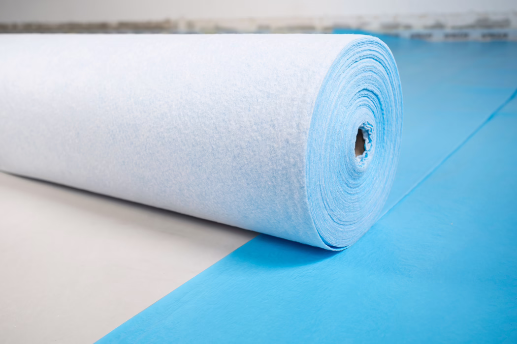 Gros plan d'un grand rouleau de tissu non tissé bleu clair ou de sous-couche posé sur une feuille bleue assortie. L'image montre la surface texturée et fibreuse et les couches en spirale enroulées autour d'un noyau en carton, posé sur un sol clair. Un éclairage doux et diffus met en valeur les fibres subtiles et la forme cylindrique ; l'arrière-plan est flou.