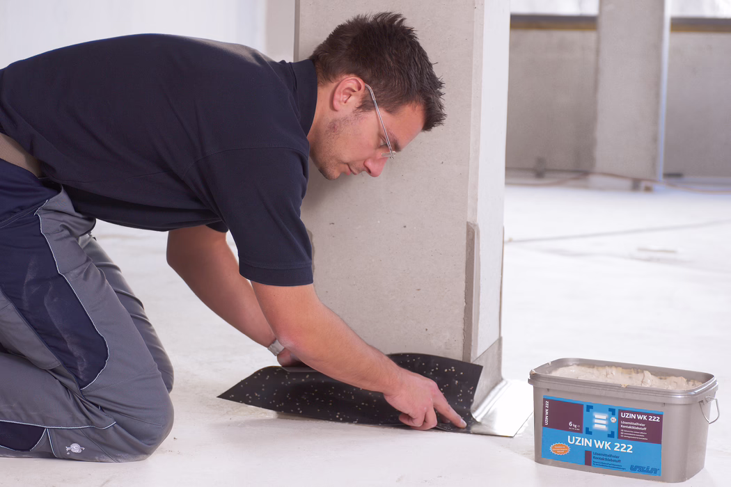 UZIN_Application_Image_WK 222_VA_3_2018-03 Un ouvrier en tenue de travail et portant des lunettes de protection est à genoux à l'intérieur d'un local, en train de poser et d'appliquer une membrane noire ou un revêtement adhésif au pied d'un mur ou d'une colonne claire. Il lisse le matériau du bout des doigts le long du bord du sol. À proximité se trouve un seau en plastique marqué 'UZIN WK 222' contenant de la colle. L'espace est un intérieur propre et inachevé avec un sol en béton ou en chape et peu d'éléments de fond.