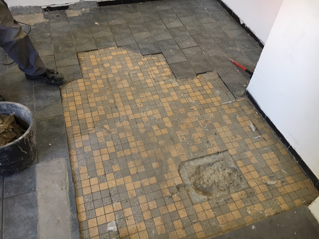 UZIN_Application_Image_UZIN NC 110_0445_ 2018-03 Sol carrelé partiellement retiré lors de travaux de rénovation : au premier plan, de petites mosaïques beige et grises usées et partiellement manquantes ; des carreaux rectangulaires foncés en périphérie ont été soulevés, révélant la colle et le sous‑plancher. À gauche, les jambes et les bottes noires d’un ouvrier sont visibles à côté d’un seau contenant des morceaux de carrelage ; à droite, un pied‑de‑biche rouge est appuyé contre le mur. Murs peints en blanc avec plinthe noire, poussière et débris éparpillés sur le sol. Photo prise en plongée depuis une faible hauteur.