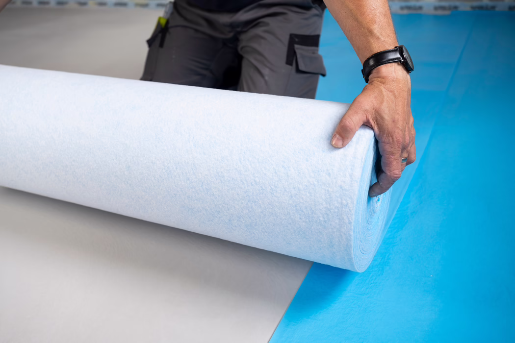 Gros plan sur la main d'un ouvrier déroulant un large rouleau d'isolant ou de sous-couche blanc et bleu pâle sur un sol bleu vif. La main droite porte une montre noire et une bague simple, saisissant le bord du rouleau ; un pantalon de travail gris avec poches et un outil clipsé sont partiellement visibles à l'arrière-plan. On distingue la texture douce et fibreuse du matériau ; la scène évoque un chantier intérieur ou une pose de revêtement de sol et la composition met en valeur la courbure du rouleau et le contraste de couleurs entre blanc, bleu pâle et bleu vif du sol.