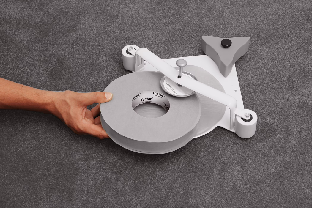UZIN_Application_Image_Toptac 35_2_2018-03 Gros plan d'une main gauche tenant un grand rouleau de ruban adhésif gris posé sur un dérouleur à profil bas sur une surface textile grise et texturée. Le dérouleur est doté d'un bras métallique blanc avec une broche centrale et une roue de coupe ronde, de deux petits rouleaux blancs à la base et d'un stabilisateur triangulaire gris avec un patin en caoutchouc noir ; le noyau en carton du ruban porte la marque imprimée 'Toptac'. Un éclairage doux et uniforme met en valeur une palette neutre de gris et de blanc. La composition centre l'attention sur le rouleau et la main en bas à gauche, montrant la couleur de la peau et les veines sans éléments permettant d'identifier la personne.