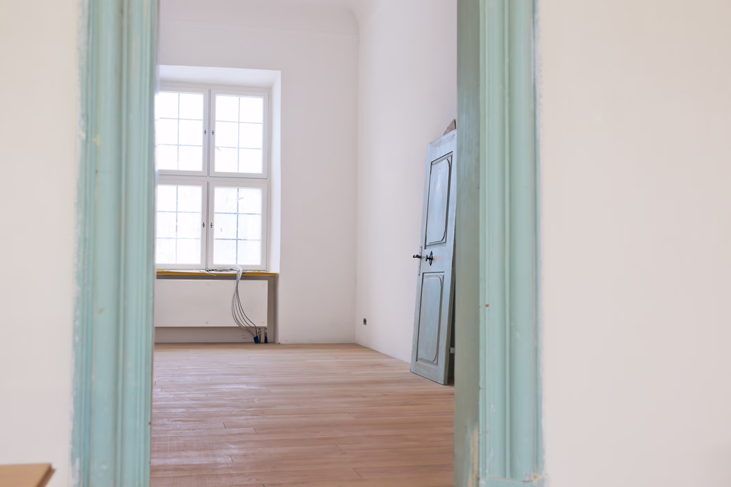 Vue intérieure à travers un chambranle peint en turquoise pâle sur une pièce vide et baignée de lumière. Une grande fenêtre à quatre carreaux, centrée sur le mur du fond, inonde l'espace d'une lumière naturelle douce. Le sol est en lames de bois claires récemment poncées, les murs sont enduits de blanc et une porte en bois bleu gris légèrement patinée est appuyée contre le mur droit. Près de la fenêtre, des tuyaux de chauffage bas et un faisceau de câbles électriques apparents reposent sur le rebord et descendent au sol, suggérant des travaux de rénovation en cours. L'ambiance générale est calme et minimale, avec des tons pastel atténués et des lignes architecturales épurées.