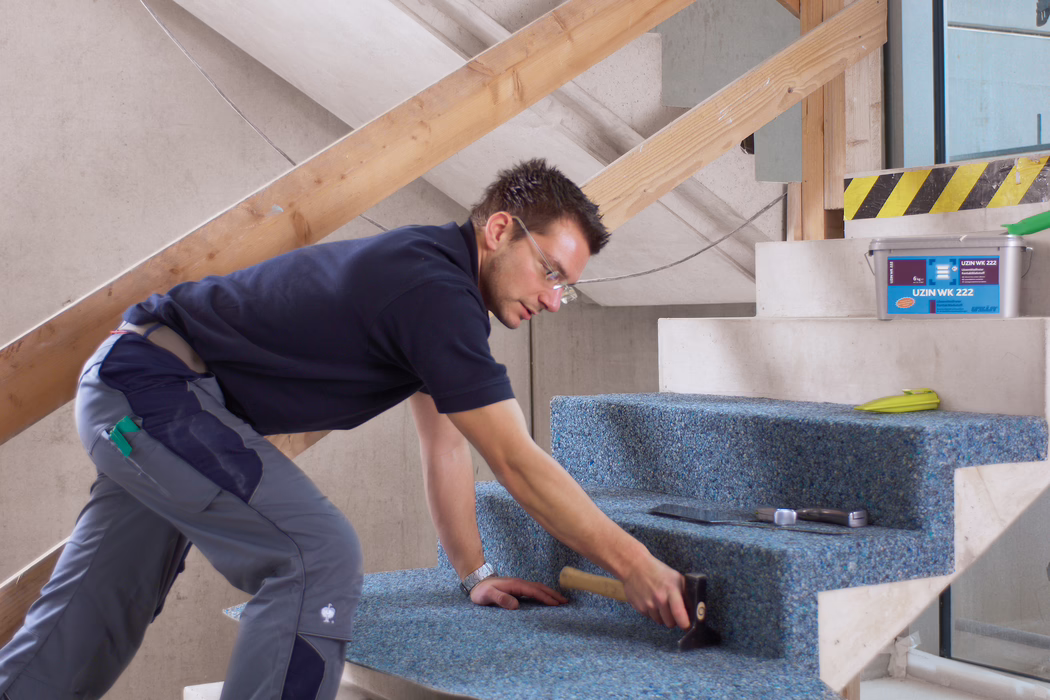 Un ouvrier pose une moquette bleue recyclée sur un escalier en béton non fini à l'intérieur d'un bâtiment en rénovation. Il est à genoux, penché en avant, porte des lunettes de protection, un polo bleu marine et un pantalon de travail gris, et utilise un maillet en caoutchouc pour bien fixer la moquette ; à proximité se trouvent une truelle, un mètre ruban et un seau en plastique marqué 'UZIN WK 222'. La scène est bien éclairée par la lumière naturelle, des planches de bois provisoires servant de rambarde sont visibles, l'accent est mis sur l'installation soignée et la texture de la moquette sur les marches.