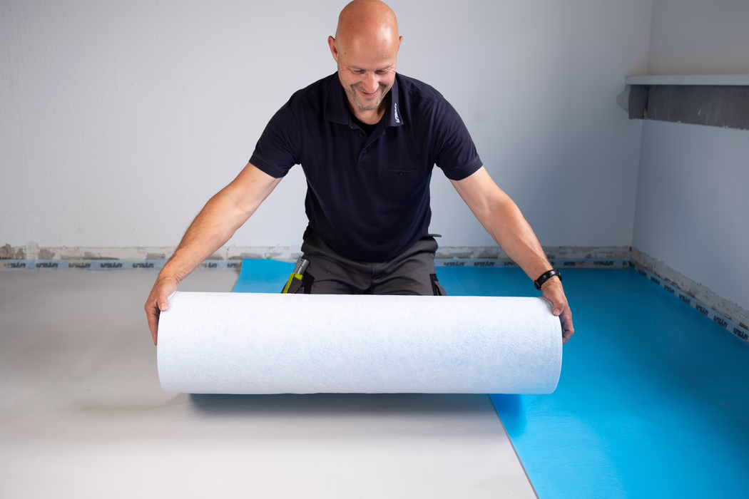 Un homme chauve d'âge moyen souriant, vêtu d'un polo sombre, déroule un rouleau blanc de sous-couche en posant un revêtement de sol sur une barrière vapeur bleue dans une pièce vide.