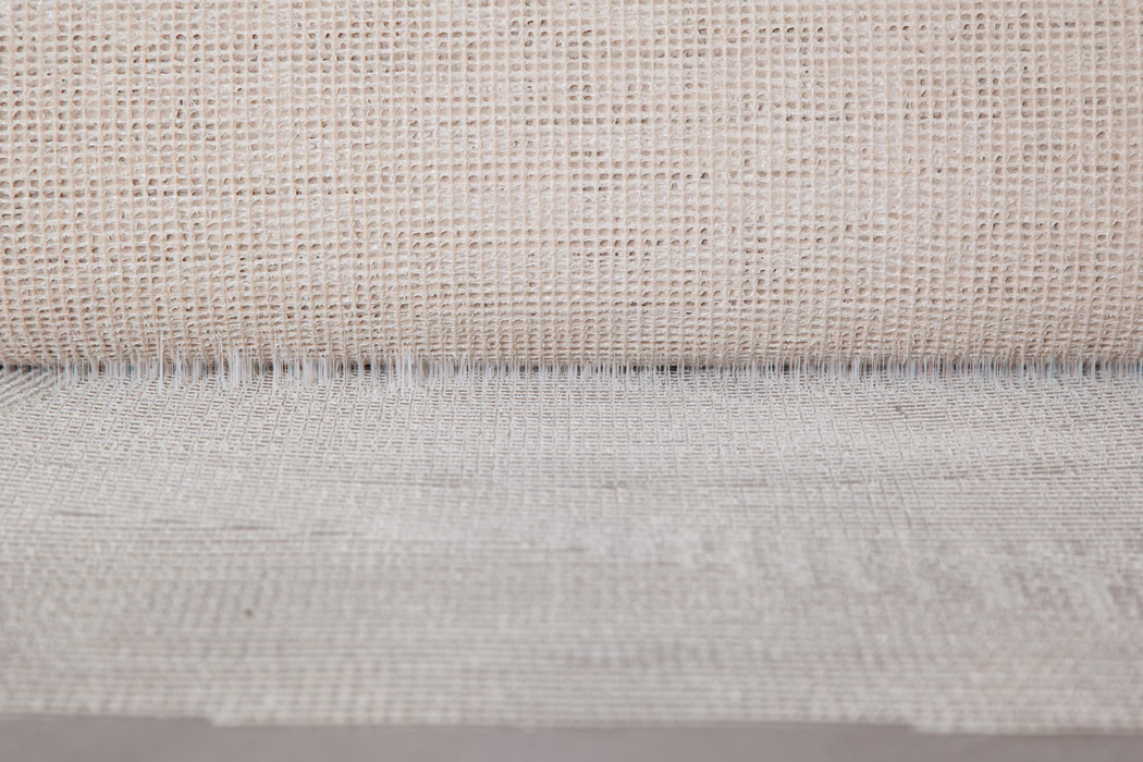 Gros plan d'un tissu tissé beige neutre à motif de tissage ouvert en grille. La partie supérieure montre un bord roulé créant une ligne d'ombre fine; des fils irréguliers et des variations de fibres naturelles donnent un aspect tactile et rustique. Un éclairage doux et uniforme met en valeur la texture et la profondeur subtile.