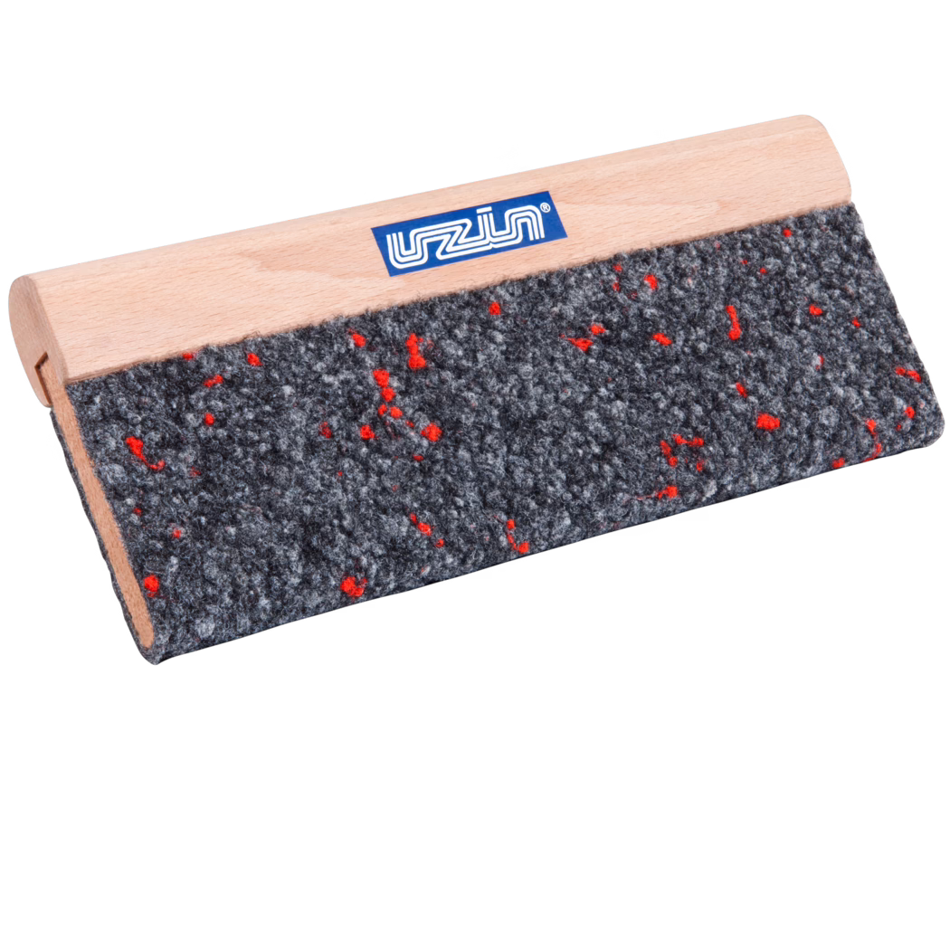 UZIN_Dummy_cork rubbing board textile covered-UZIN_2014-01 Gros plan d'un outil rectangulaire avec une poignée en bois clair et un patin en feutre gris grossier parsemé de petites fibres rouges ; une petite étiquette de marque bleue est visible sur la poignée en hêtre lisse. L'outil est photographié en angle sur fond blanc, mettant en valeur la texture du feutre et le grain fin du bois.